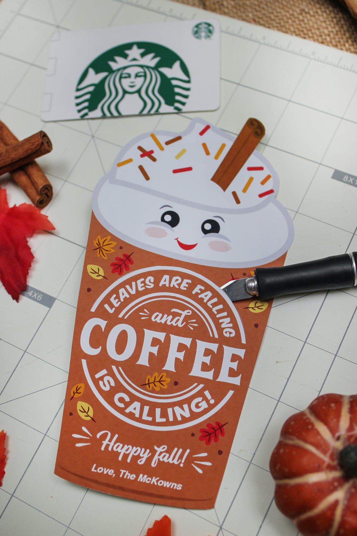 A fall-themed design card with a smiling coffee cup illustration, topped with whipped cream, colorful sprinkles, and a cinnamon stick, displayed on a crafting grid. The card reads, 'Leaves are falling and coffee is calling! Happy fall! Love, The McKowns.' Surrounding items include a Starbucks gift card in the top left, cinnamon sticks, red fabric leaves, a small pumpkin, and a black craft knife resting on the card.