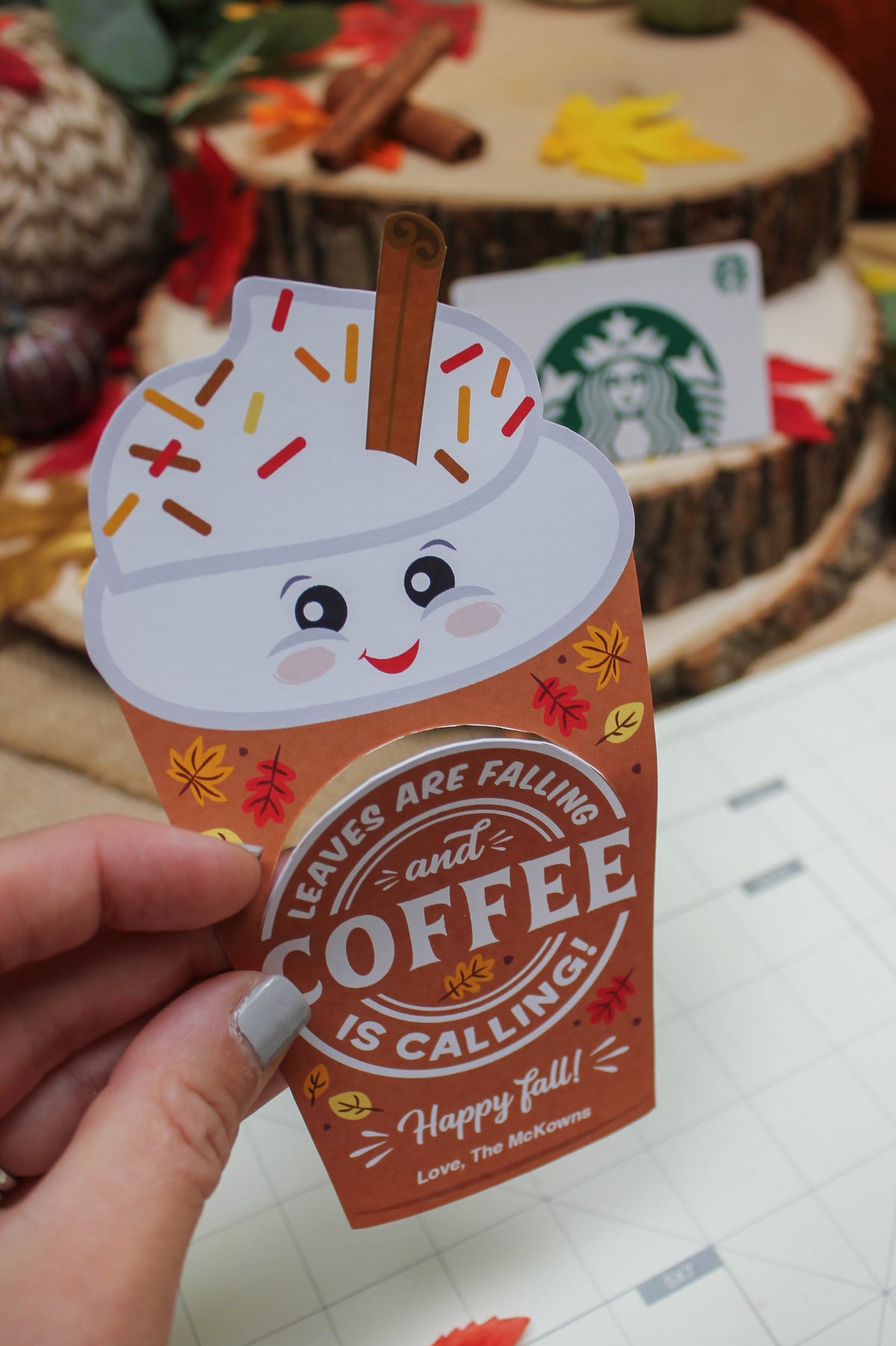 A close-up of a hand holding a fall-themed card cutout shaped like a smiling coffee cup with whipped cream, colorful sprinkles, and a cinnamon stick. The card reads, 'Leaves are falling and coffee is calling! Happy fall! Love, The McKowns.' The background includes a wood slice display with fall decorations such as cinnamon sticks, fabric leaves, and a partially visible Starbucks gift card.